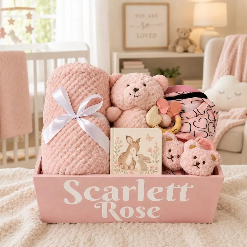 A personalized baby gift basket labeled 'Scarlett Rose' filled with soft pink items, including a textured blanket tied with a white ribbon, a teddy bear, a patterned pouch, plush toys, and a deer-themed card, set in a cozy nursery.